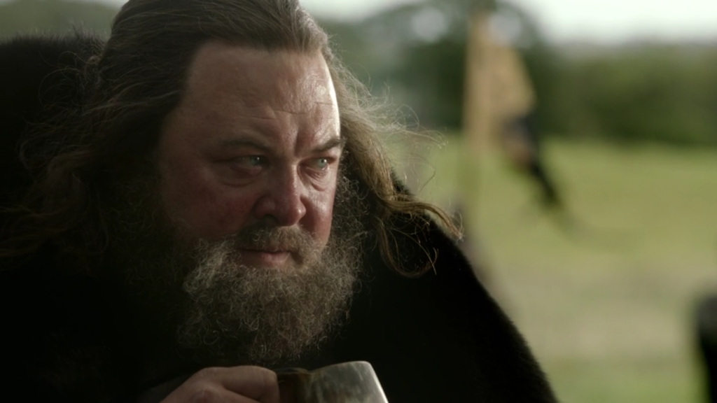 Closeup of King Robert in conversation, holding a mug.